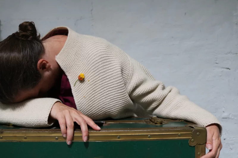 Schauspielerin Beate Krist lehnt auf einem Koffer mit dem Kopf in den Arm versteckt