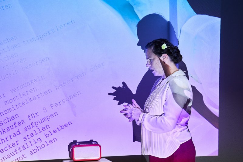 Schauspielerin Beate Krist im Monolog. Sie steht in der Projektion einer Liste vor einem Radio.