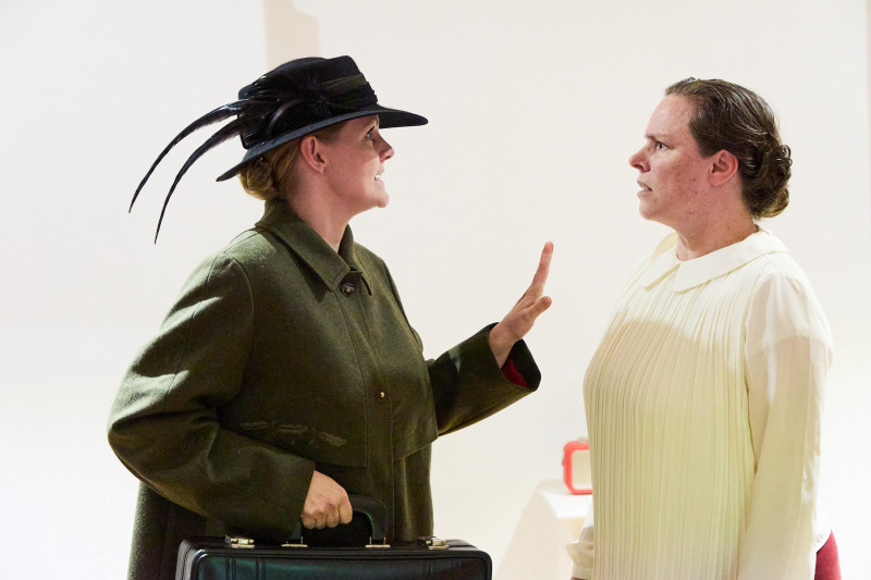 Schauspielerinnen im Dialog. Die linke trägt einen braunen Mantel, einen schwarzen Koffer und einen Hut mit Federschmuck. Sie hält die Hand abwehrend der anderen entgegen. Die Schauspielerin rechts schaut als würde sie ungerecht behandelt.