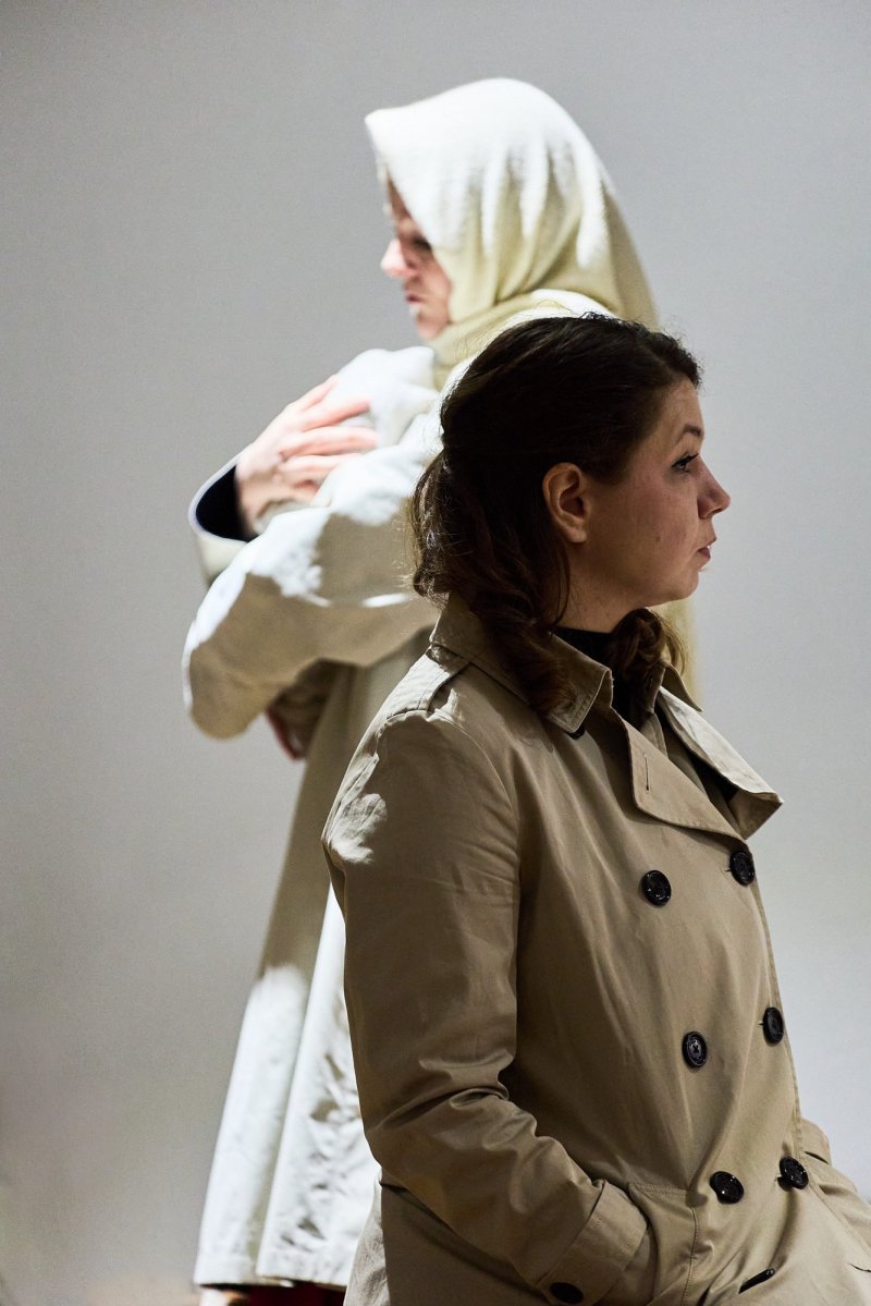 Szenenfoto. Zwei Frauen stehen in entgegengesetzer Richtung hintereinander in Mantel, es wirkt wie eine Straßenszene. Die hintere Frau trägt ein warmes Kopftuch und im Arm ein Baby.