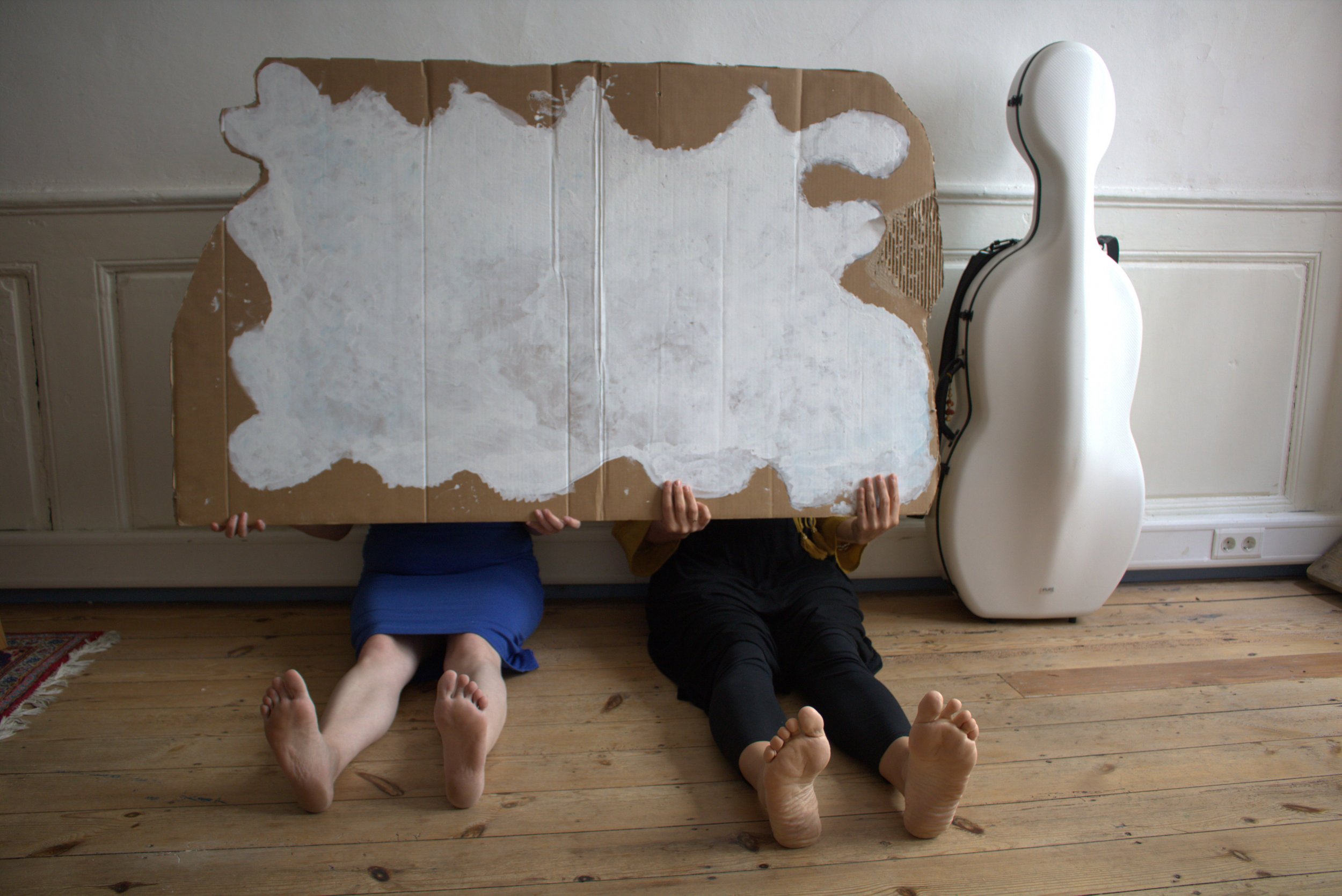Zwei Frauen sitzen auf dem Boden und halten sich eine Wellpappe mit einer aufgemalten Wolke vor den Oberkörper/Gesicht. Die Personen sind nicht sichtbar. Neben ihnen steht ein weißer Cello-Koffer.