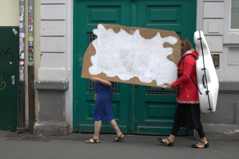 Zwei Frauen tragen auf der Straße eine große Wellpappe mit einer aufgemalten Wolke. Die hintere Frau trägt auf dem Rücken einen weißen Cellokoffer.