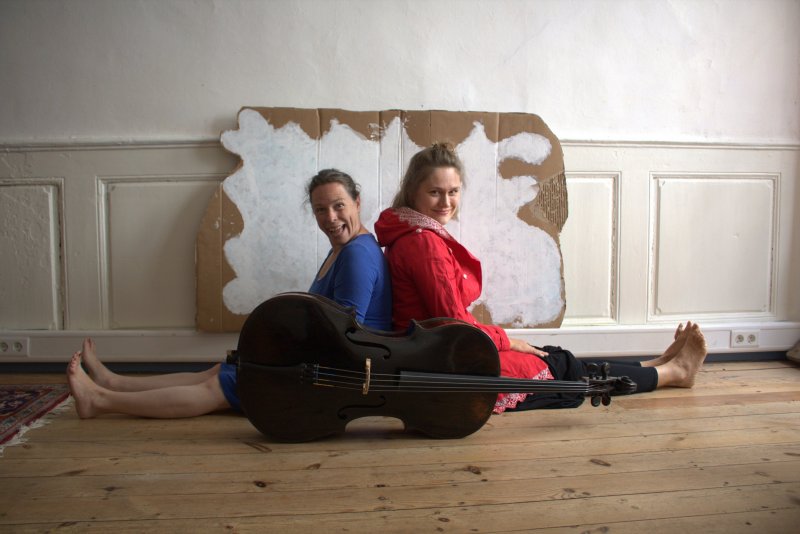 Zwei Frauen sitzen auf dem Boden Rücken an Rücken und blicken seitlich in die Kamera. Hinter Ihnen an der Wand lehnt eine Wellpappe mit einer aufgemalten Wolke. Vor ihnen liegt ein Cello.