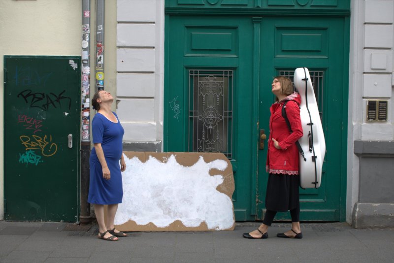 Zwei Frauen stehen vor einem Hauseingang auf dem Bürgersteig und schauen in den Himmel. Hinter ihnen an der Wand lehnt eine Wellpappe mit einer aufgemalten Wolke. Die rechte Frau trägt einen weißen Cellokoffer auf dem Rücken.