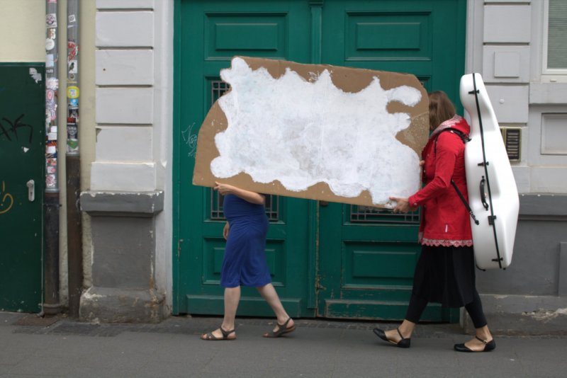 Zwei Frauen tragen auf der Straße eine große Wellpappe mit einer aufgemalten Wolke. Die hintere Frau trägt auf dem Rücken einen weißen Cellokoffer.