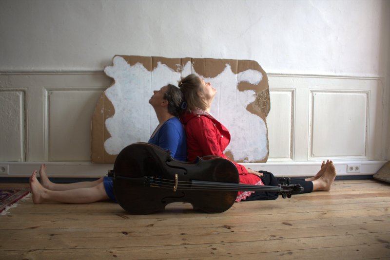 Zwei Frauen sitzen auf dem Boden Rücken an Rücken und schauen an die Decke. Hinter Ihnen an der Wand lehnt eine Wellpappe mit einer aufgemalten Wolke. Vor ihnen liegt ein Cello.
