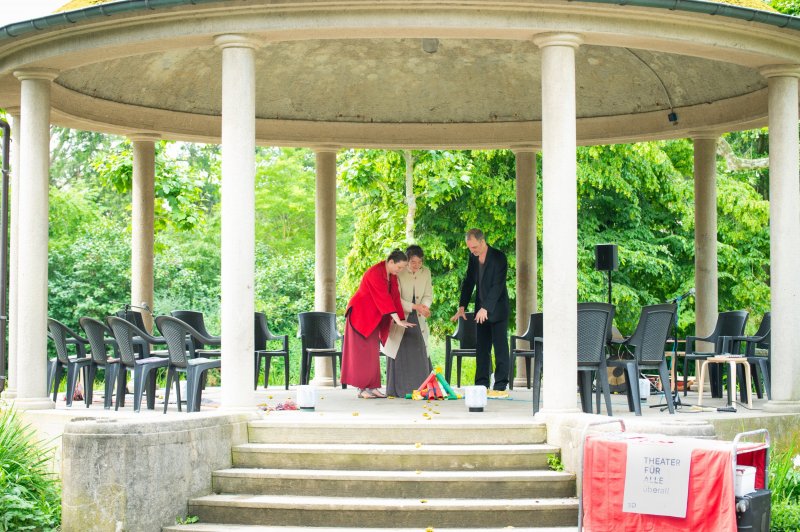 Drei Schuspieler:innen stehen um ein „Lagerfeuer“ aus Schriftrollen in einem Kurpavillon