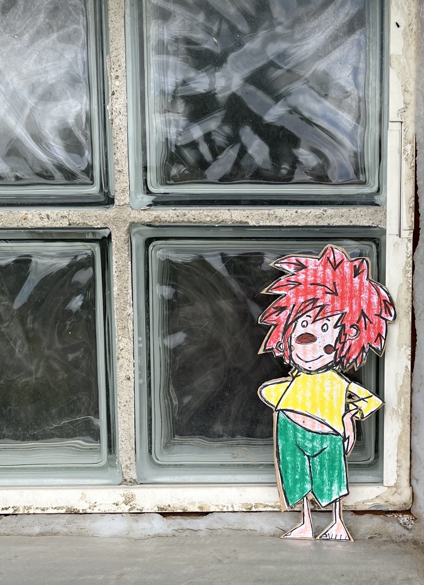Ein auf Karton gemalter und ausgeschnittener Pumuckl steht an einem Fenster aus Glasbausteinen