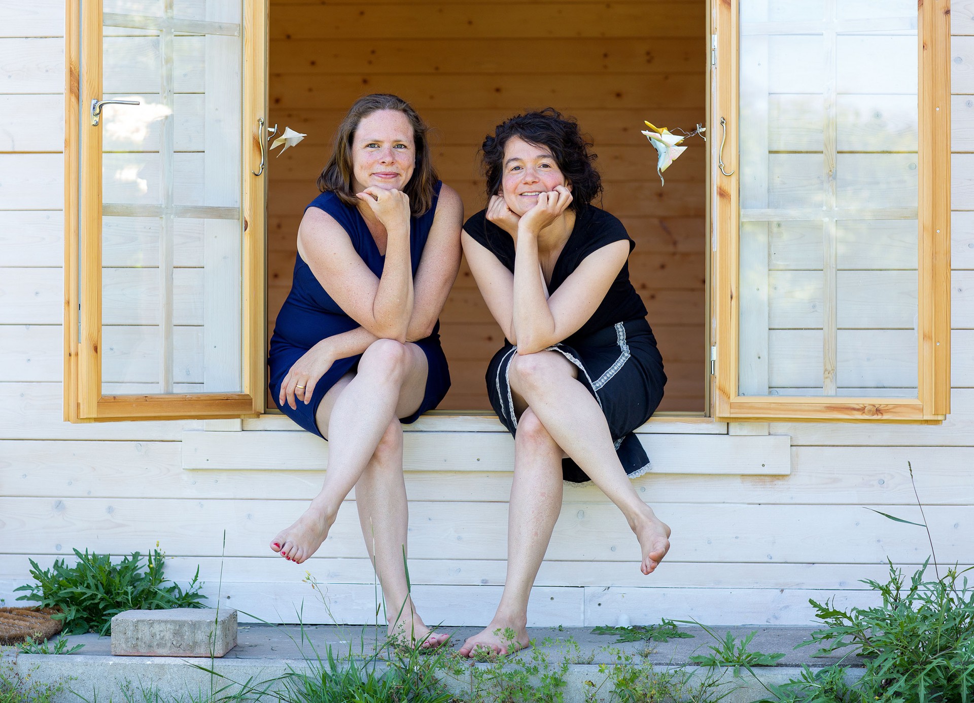 Beate Krist und Manuela Pirozzi sitzen im Fenster einer sommerlichen Holzhütte